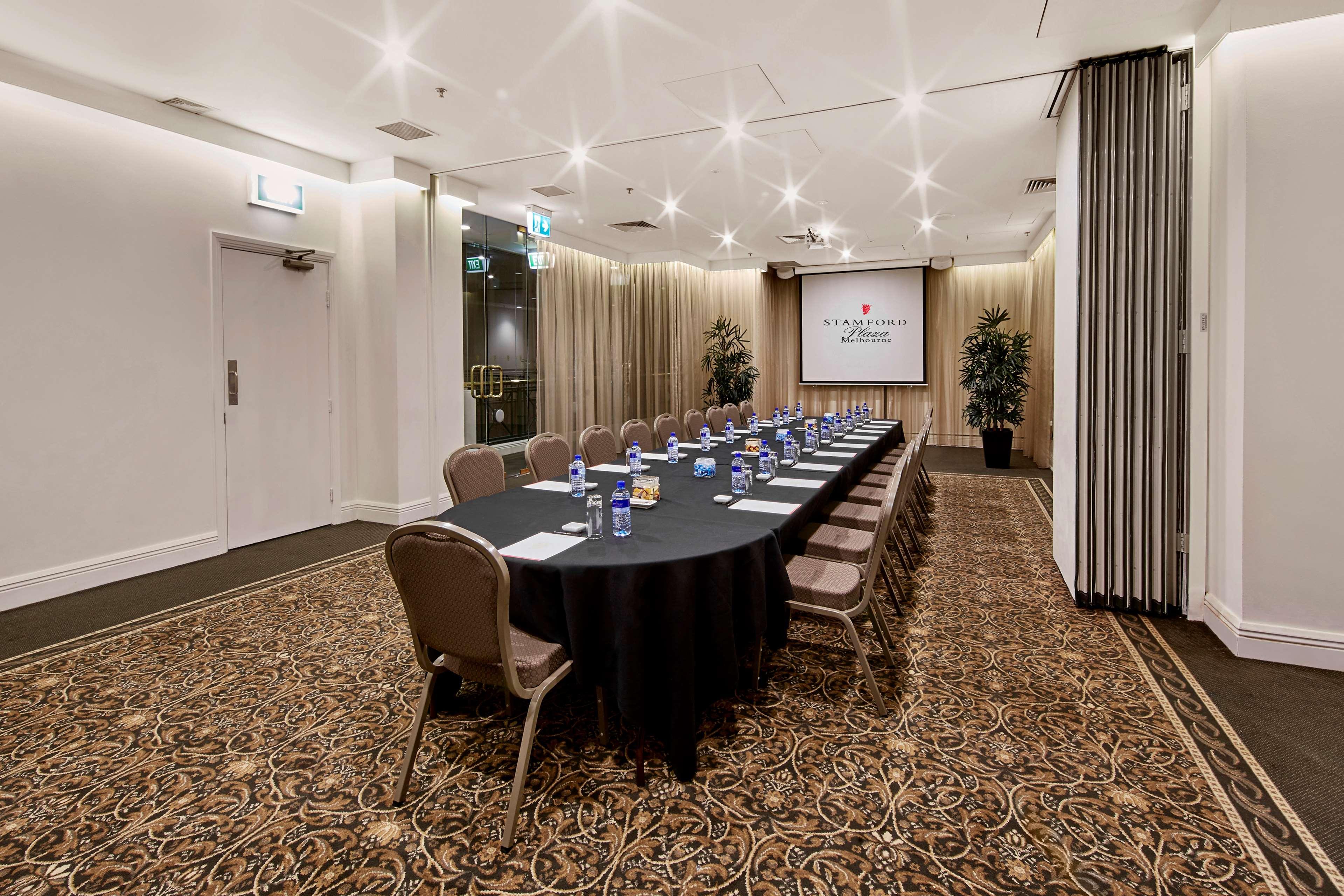 스탬포드 플라자 멜버른 호텔 멜번 외부 사진 The photo shows a meeting room set up for a conference or presentation. There is a long, rectangular table covered with a black tablecloth, surrounded by chairs. Each seat has a water bottle placed in front of it. At the front of the room, there is a