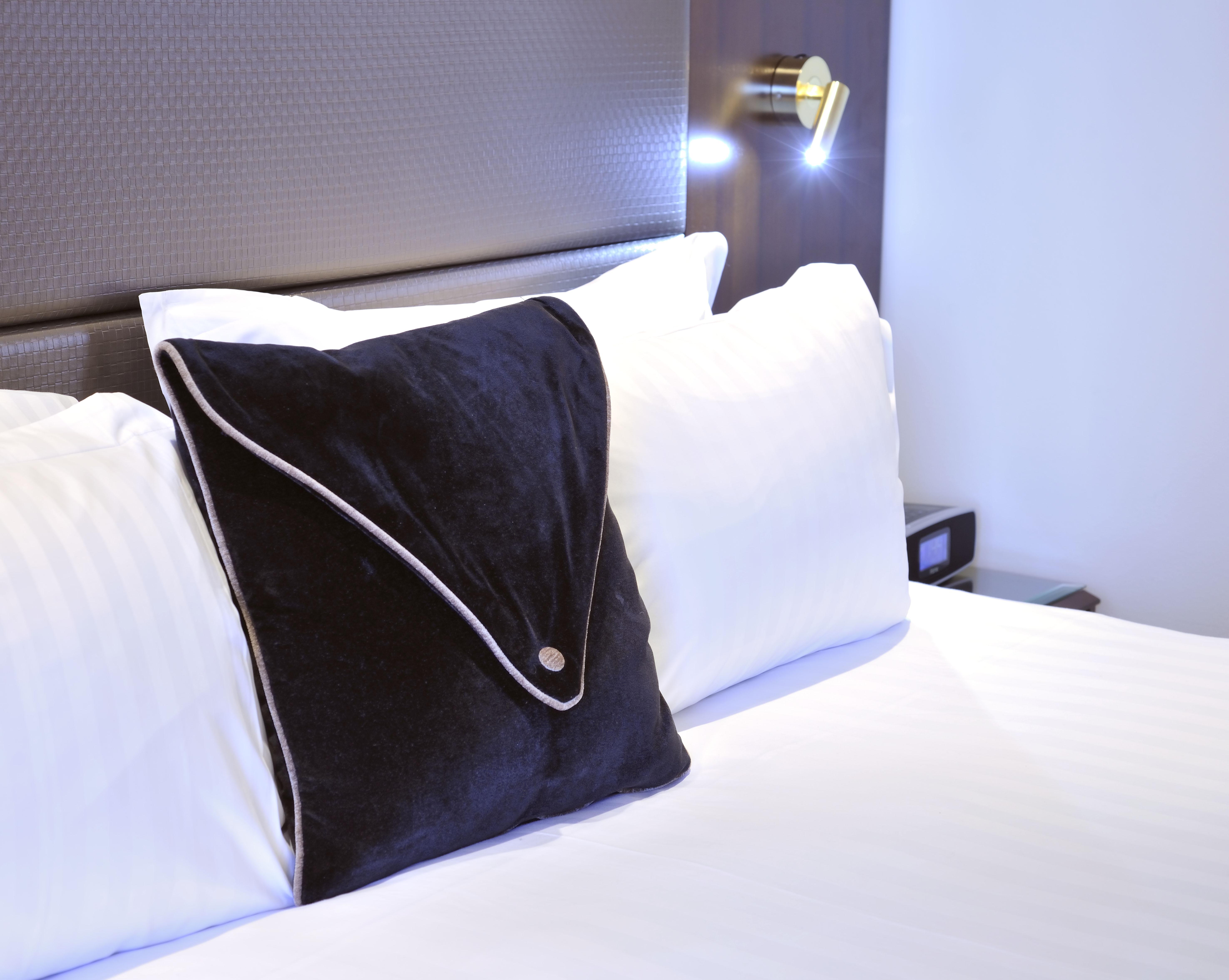 스탬포드 플라자 멜버른 호텔 멜번 외부 사진 The photo shows a neatly arranged bed with white linens. There are several pillows, with one black pillow featuring a distinctive design on top of the white pillows. The headboard of the bed is visible, along with a stylish wall-mounted light fixture