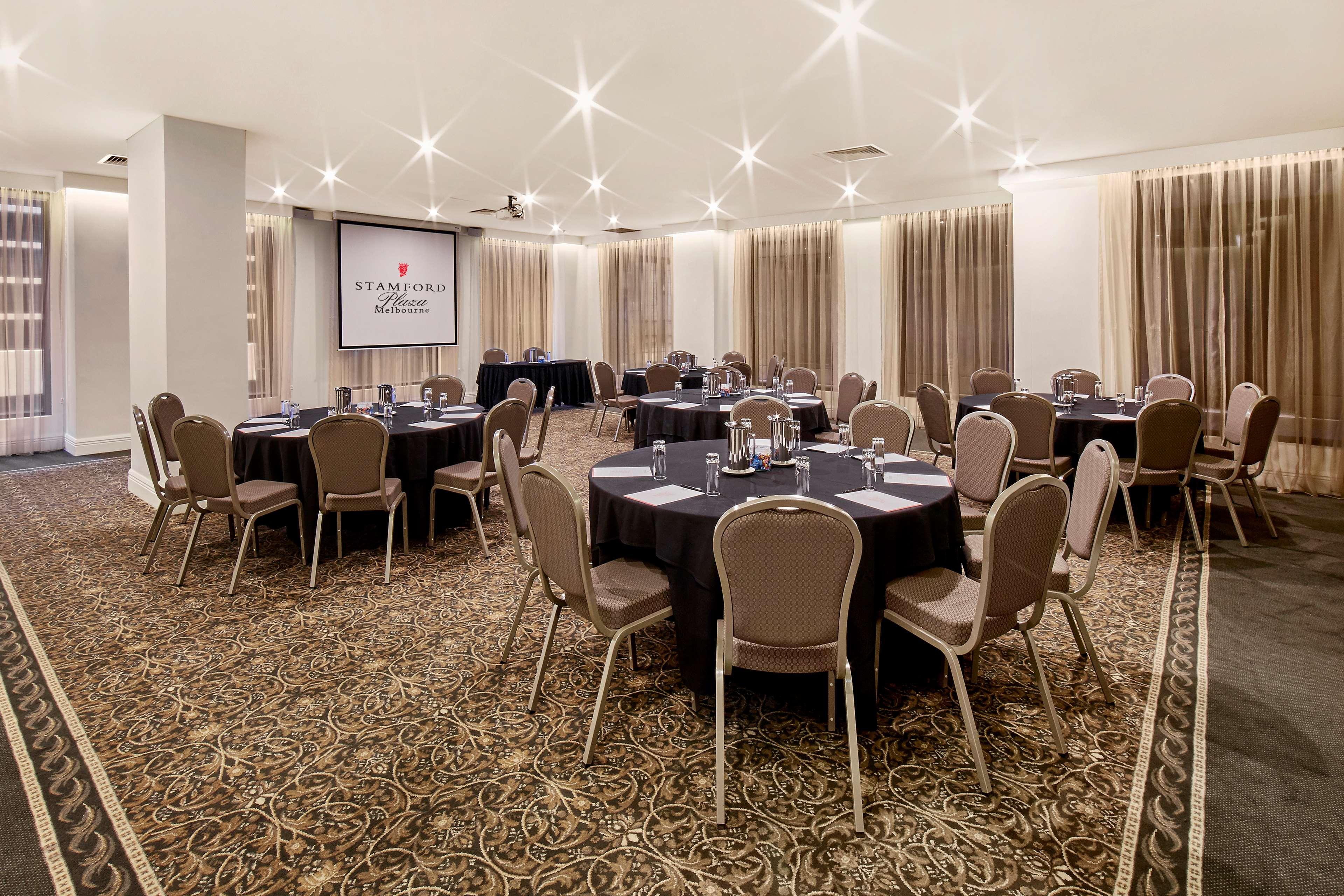 스탬포드 플라자 멜버른 호텔 멜번 외부 사진 The photo shows a well-lit event space set up for a gathering or meeting. There are several round tables covered with black tablecloths, each surrounded by beige chairs. In the background, a large screen displays a logo or branding, likely related to