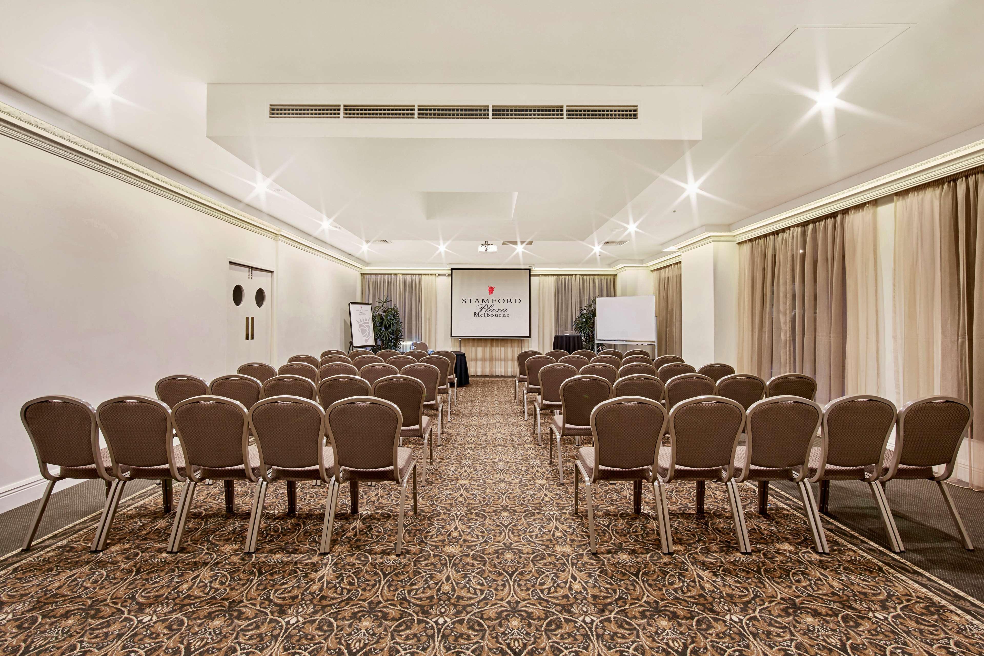 스탬포드 플라자 멜버른 호텔 멜번 외부 사진 The photo shows a conference room set up for an event. It features several rows of beige chairs arranged facing a central presentation area. At the front, there is a screen displaying a logo or title, possibly for a conference or meeting. The walls h