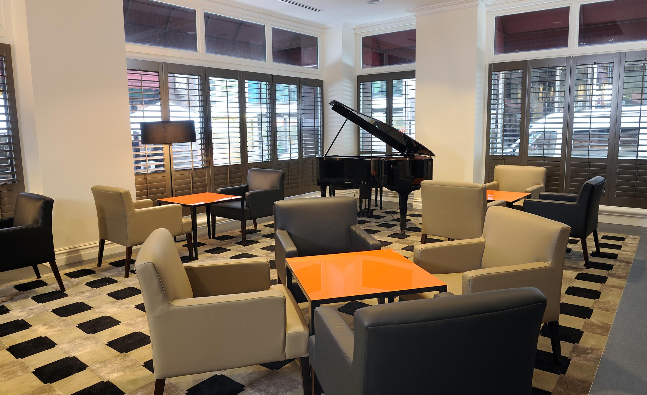 스탬포드 플라자 멜버른 호텔 멜번 외부 사진 The photo shows a stylish indoor lounge area. It features several light gray armchairs arranged around orange square tables. In the corner, there is a black grand piano, adding an elegant touch to the space. The room has large windows with wooden shu