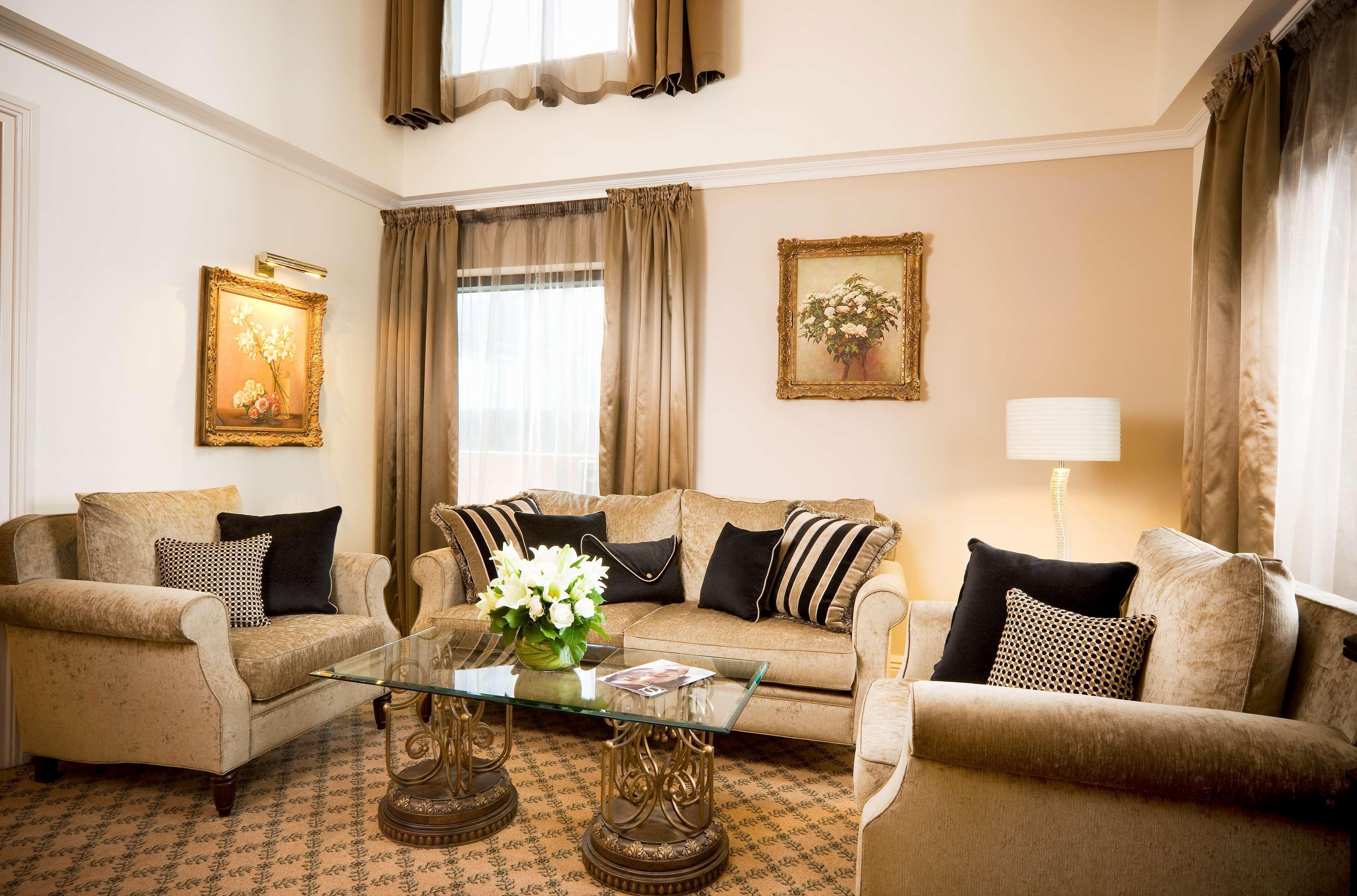 스탬포드 플라자 멜버른 호텔 멜번 객실 사진 The photo depicts a stylish living room with a warm and inviting aesthetic. It features a set of comfortable beige sofas adorned with decorative pillows in black and patterned designs. There is a glass coffee table at the center, supported by elegant