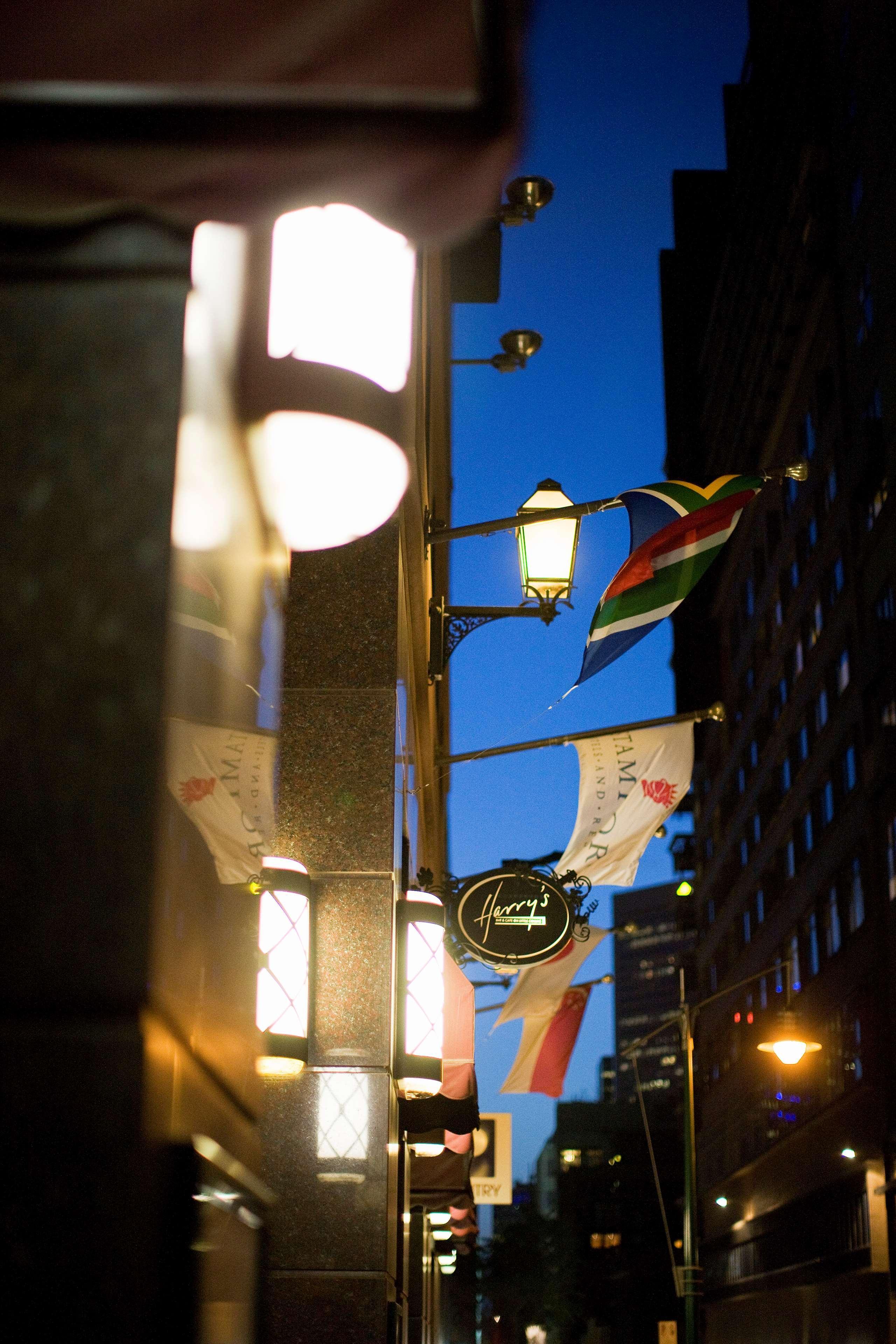 스탬포드 플라자 멜버른 호텔 멜번 외부 사진 The photo features a street view of a building at dusk. Prominently displayed are several flags, including a South African flag, which are fluttering in the wind. There is a sign that reads "Harry's," likely indicating the name of a business or estab