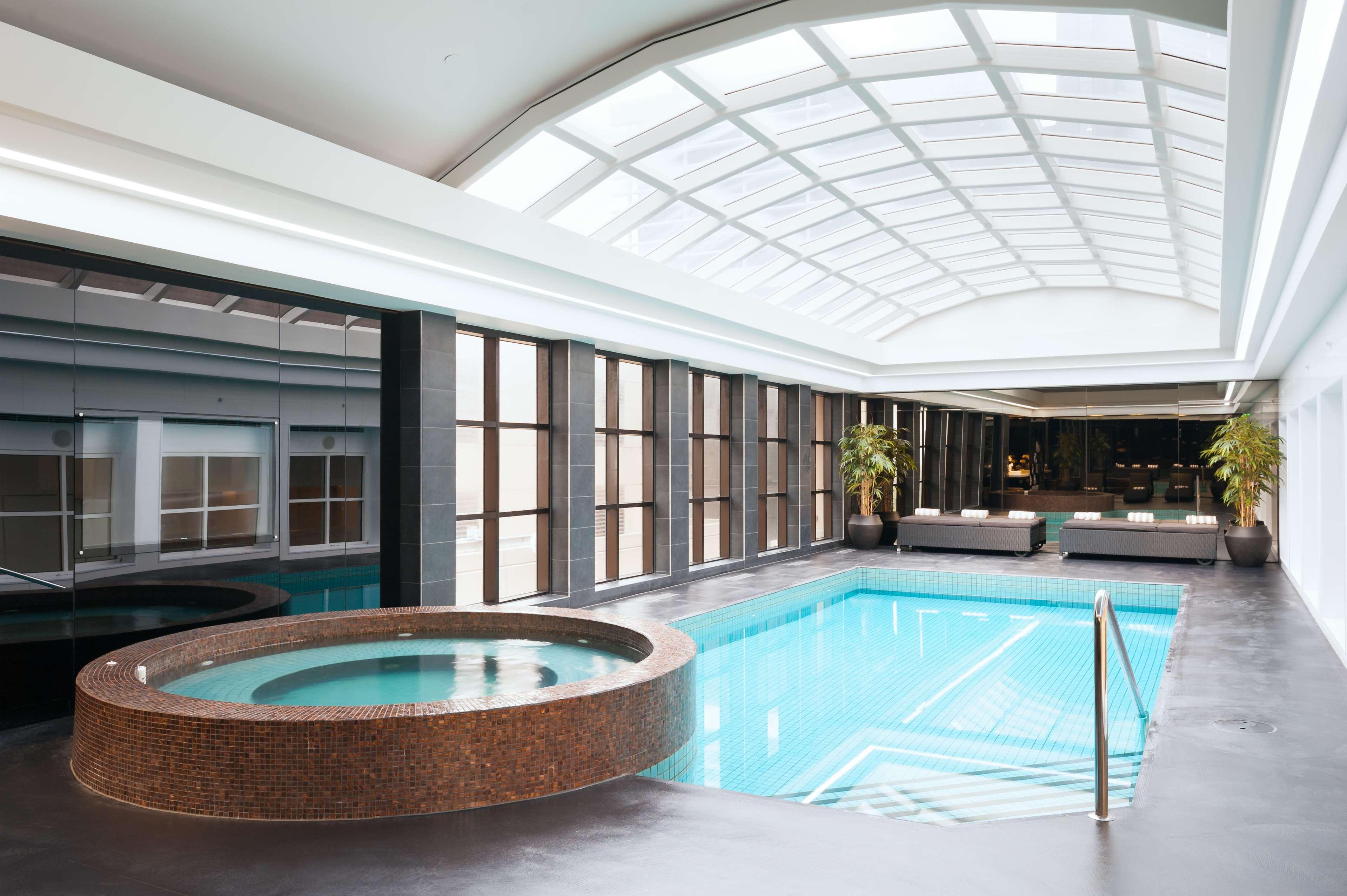 스탬포드 플라자 멜버른 호텔 멜번 외부 사진 The photo shows an indoor swimming pool area. The pool is rectangular and has clear blue water, surrounded by a sleek, modern design. On one side, there is a round hot tub made of dark stone. Large windows line the walls, allowing natural light to fi