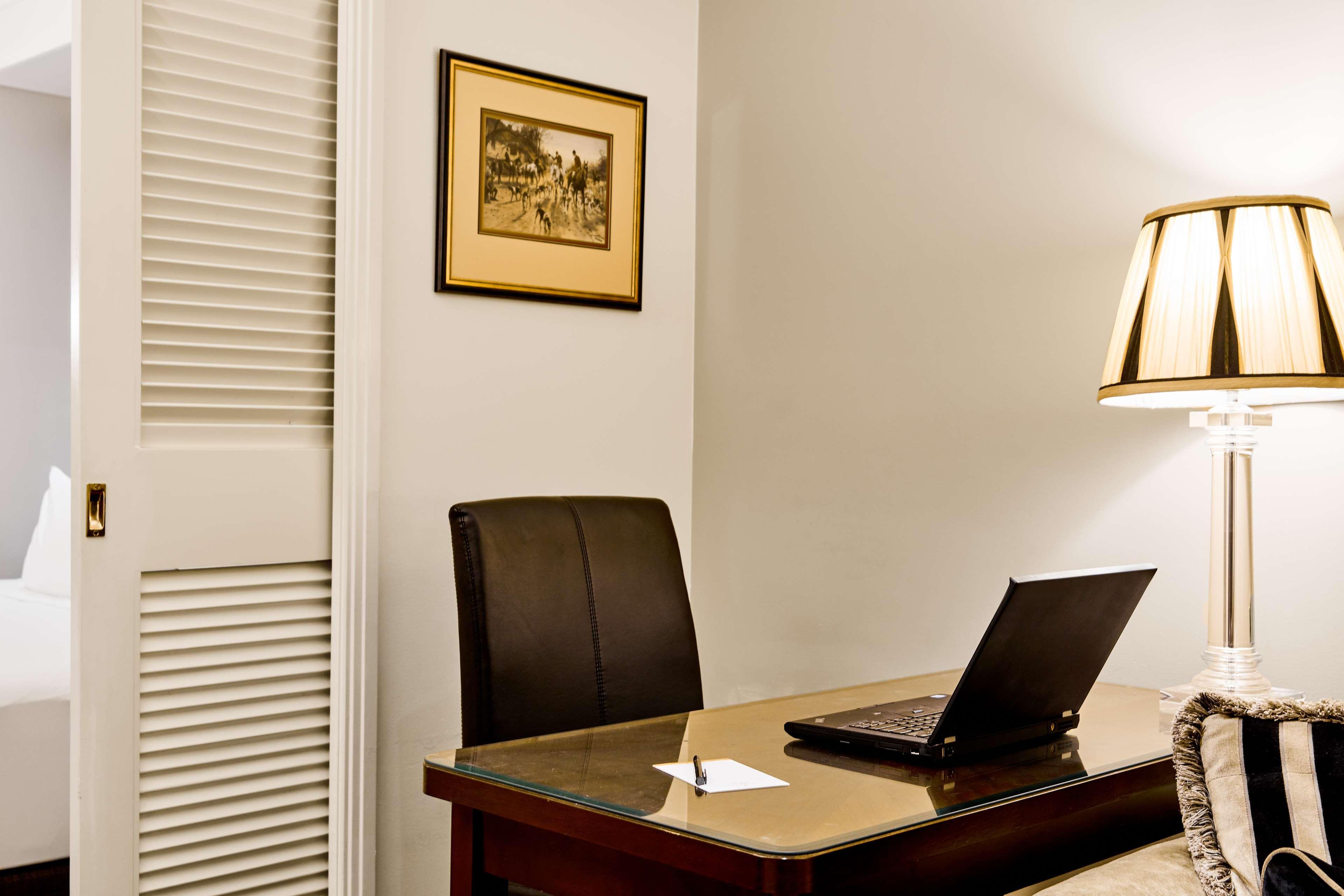 스탬포드 플라자 멜버른 호텔 멜번 외부 사진 The photo shows a small office or workspace. There is a desk with a glass surface, and on it sits a laptop computer. A piece of paper or notecard is also placed on the desk. Behind the desk is a black office chair. A wall lamp with a striped shade is