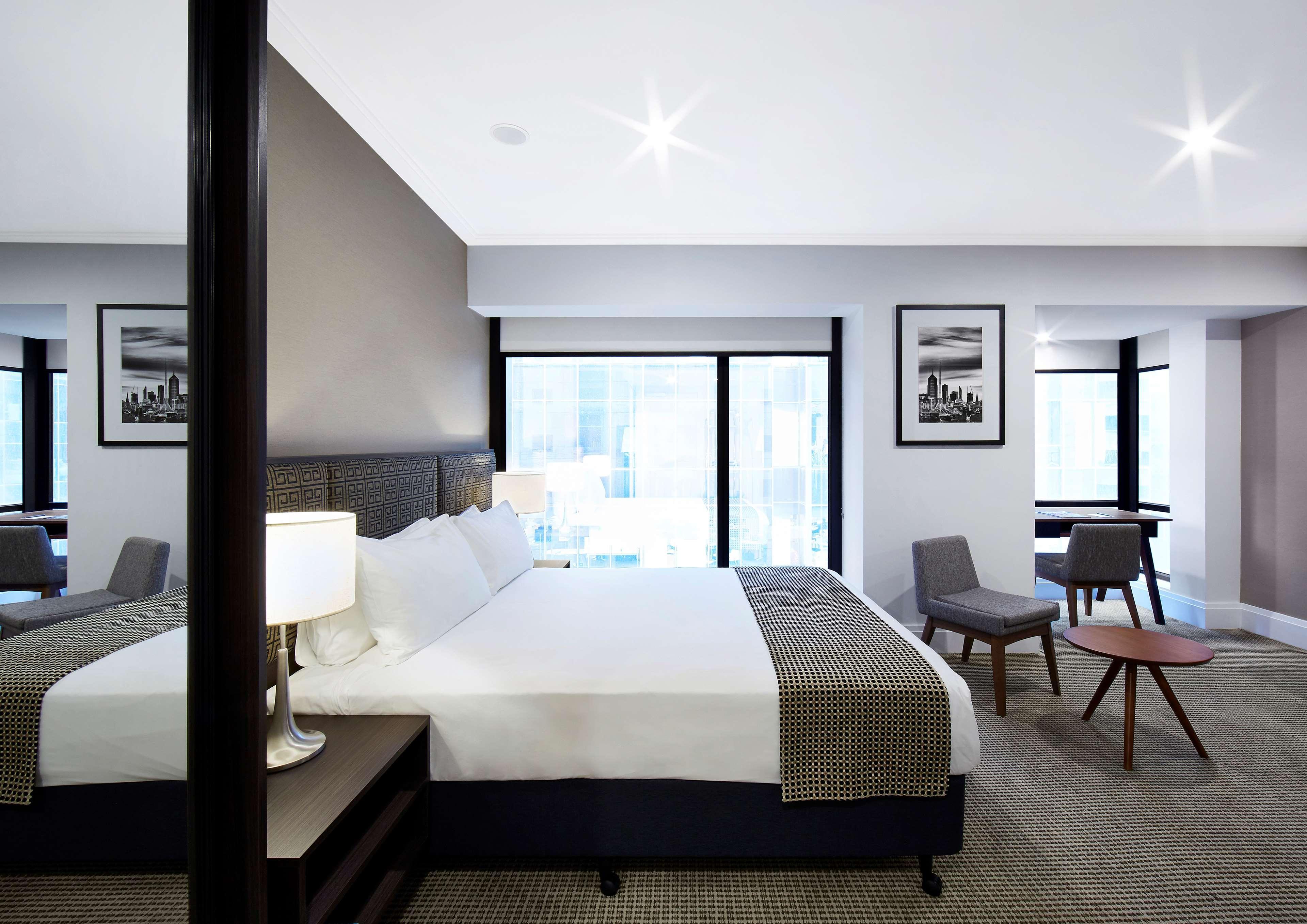 스탬포드 플라자 멜버른 호텔 멜번 외부 사진 The image shows a modern hotel room featuring a large bed with white linens and a gray throw blanket. There are two windows that allow natural light to enter the space, and the walls are painted in a light color. In addition to the bed, there are two
