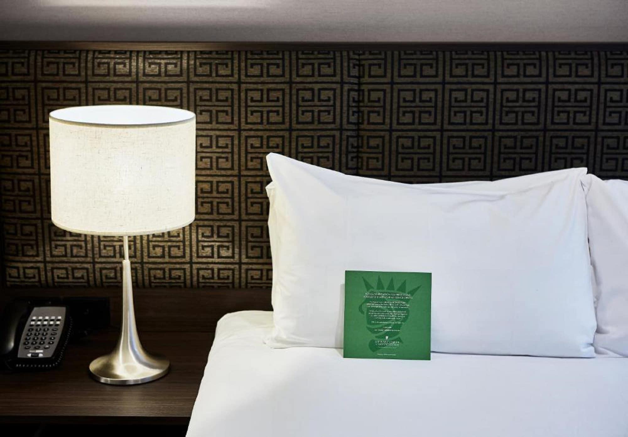 스탬포드 플라자 멜버른 호텔 멜번 외부 사진 The photo shows a neatly made bed with a white pillow and a green item placed on top of it. Next to the bed, there is a stylish table lamp with a lampshade that appears to be white. In the background, you can see a patterned wall, which has a geometr
