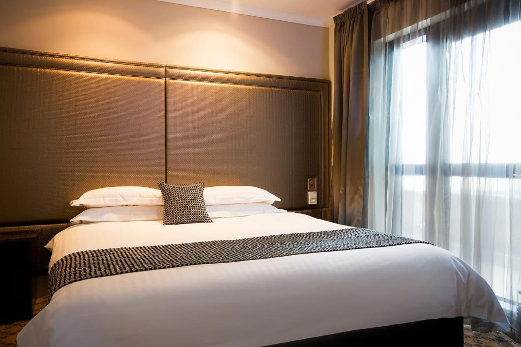 스탬포드 플라자 멜버른 호텔 멜번 외부 사진 The photo shows a modern bedroom featuring a large bed with a neatly arranged white comforter and decorative pillows. The headboard is upholstered in a dark material, and there is soft lighting above it. On one side of the bed, there’s a small decora
