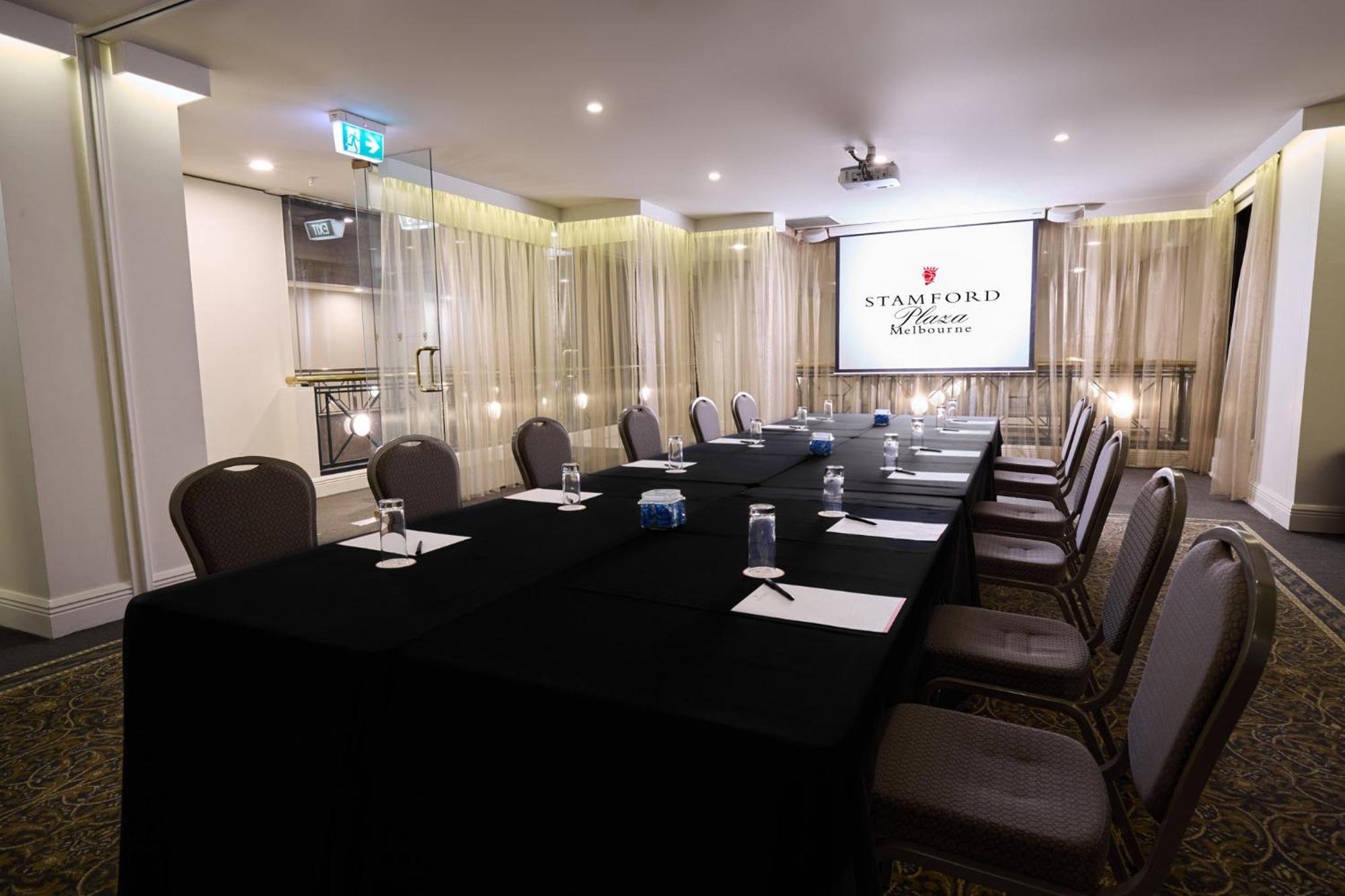 스탬포드 플라자 멜버른 호텔 멜번 외부 사진 The photo shows a well-appointed conference room designed for meetings or presentations. There is a long rectangular table covered with a black tablecloth, surrounded by upholstered chairs. On the table, there are several folded pieces of paper and g