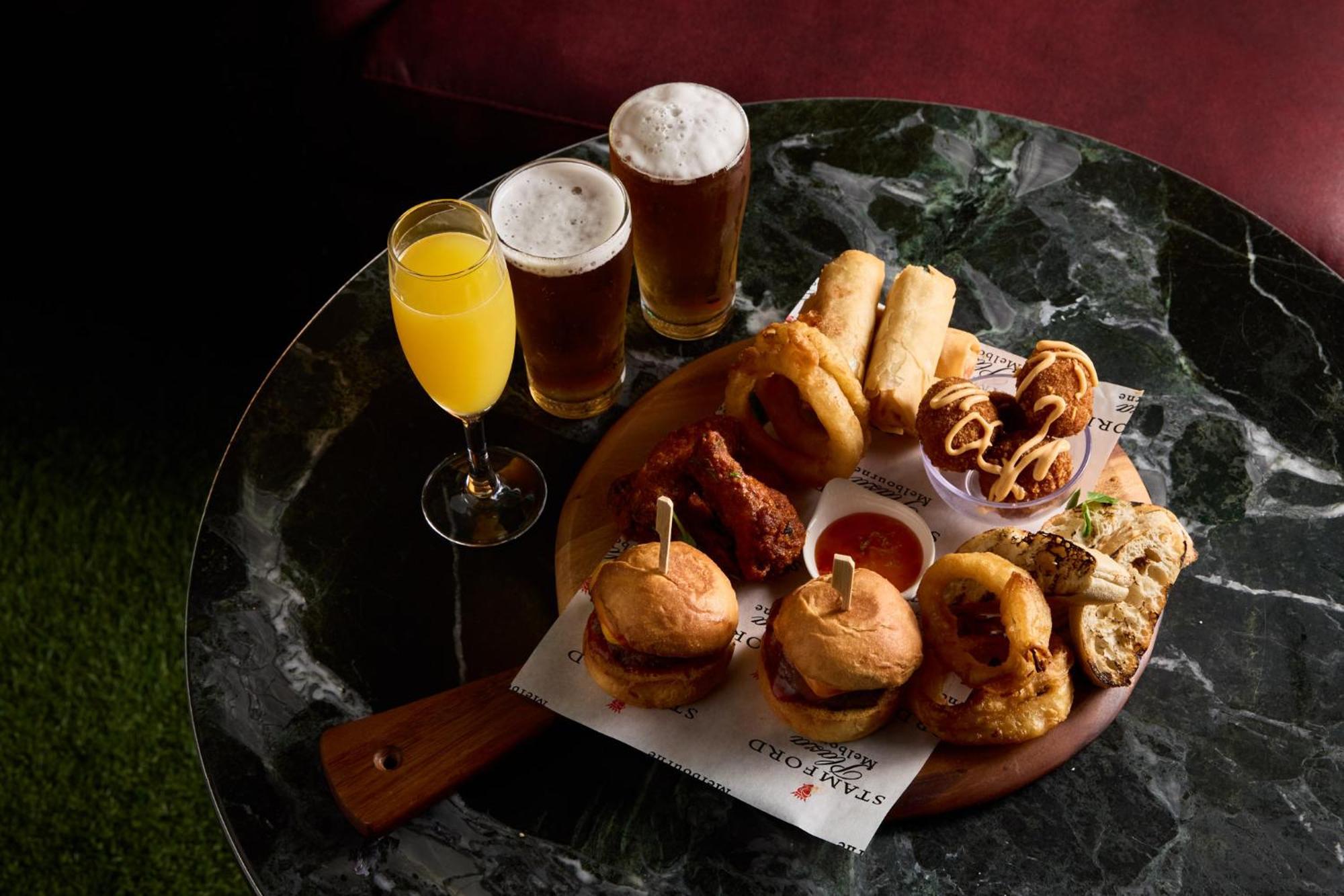 스탬포드 플라자 멜버른 호텔 멜번 외부 사진 The photo displays a variety of food and drinks arranged on a marble table. There are small burgers garnished with skewers, crispy chicken pieces, and deep-fried snacks. Additionally, there are two glasses of beer and a cocktail, likely a mimosa, pre