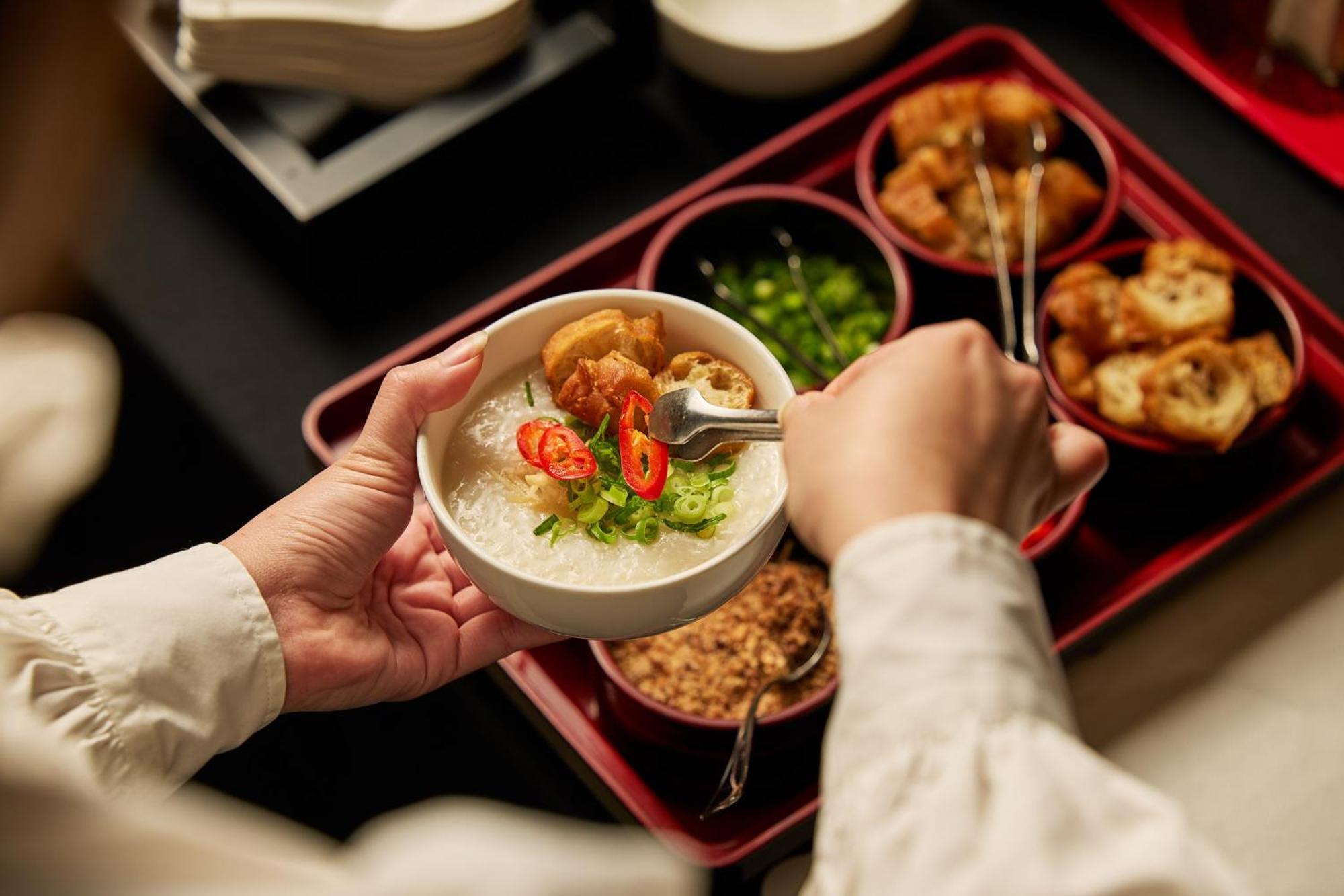 스탬포드 플라자 멜버른 호텔 멜번 외부 사진 The photo shows a person serving food from a tray filled with various dishes. In one hand, they are holding a bowl filled with a creamy, likely rice-based dish topped with slices of chili and possibly fried tofu or other toppings. Surrounding the bow