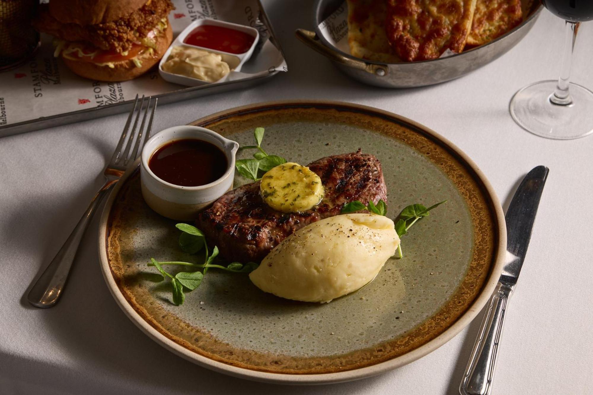 스탬포드 플라자 멜버른 호텔 멜번 외부 사진 The photo shows a well-presented plate featuring a serving of steak, topped with a dollop of herb butter and surrounded by fresh sprouts or microgreens. There is a smooth portion of mashed potatoes beside the steak, and a small bowl of sauce, likely 