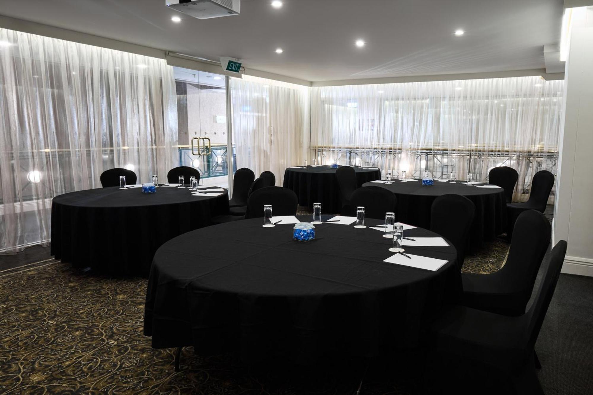 스탬포드 플라자 멜버른 호텔 멜번 외부 사진 The photo shows a meeting or conference room set up with several round tables covered in black tablecloths. Each table is accompanied by black chairs. There are some white notepads and pens placed on the tables, and a small decorative item, possibly 